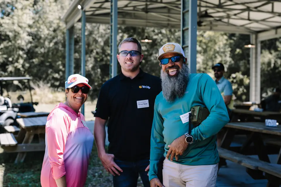 Tri-City Electrical Contractors Three people smiling at an outdoor gathering. One wears a pink hat, another wears sunglasses and a black shirt, and the third wears a beige cap and blue sunglasses. They are standing in front of wooden picnic tables under a pavilion.