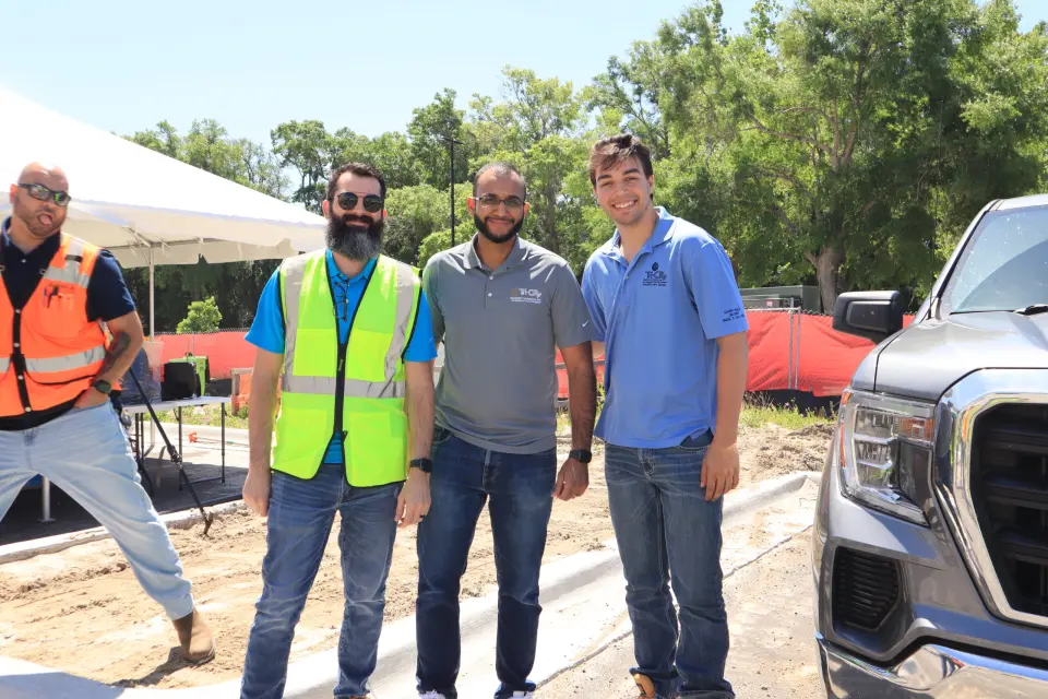 Tri-City Electrical Contractors Three men stand smiling at a construction site. One wears a yellow safety vest, and another a blue polo. Trees, a white canopy, and a parked vehicle are in the background. A fourth man in an orange vest is on the left edge of the photo.