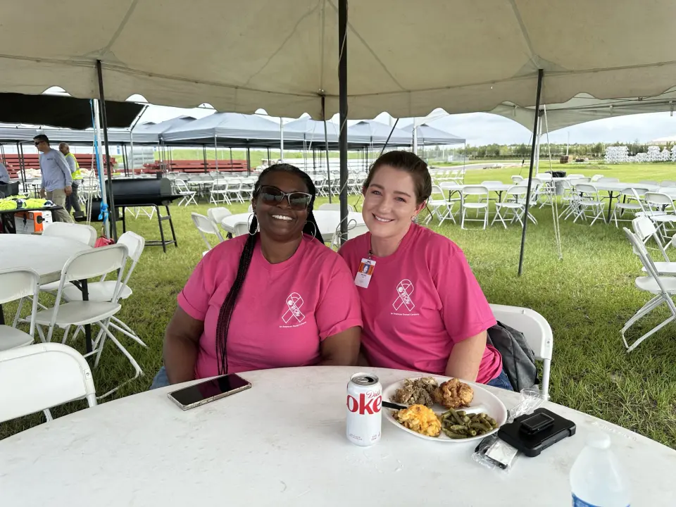 Tri-City Electrical Contractors Two people in pink shirts sit at a round table under a tent, smiling. One has a soda can, the other has a plate of food. White folding chairs and tables are in the background on green grass.
