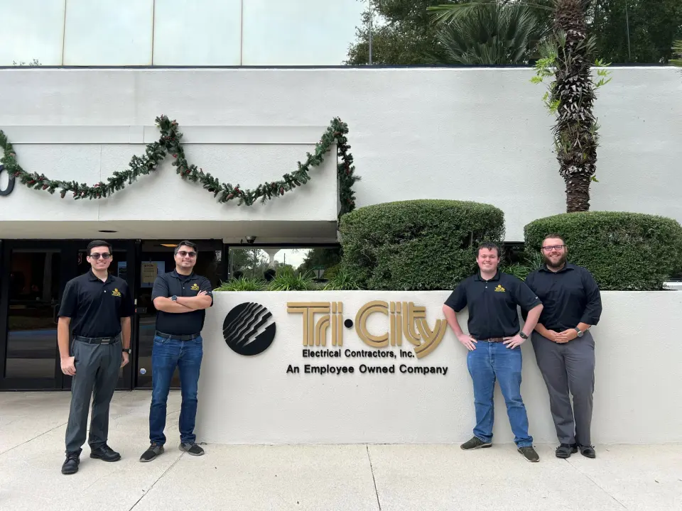 Tri-City Electrical Contractors Four men stand smiling in front of a building with the sign Tri-City Electrical Contractors, Inc. The building is decorated with greenery. They are wearing dark shirts and jeans.