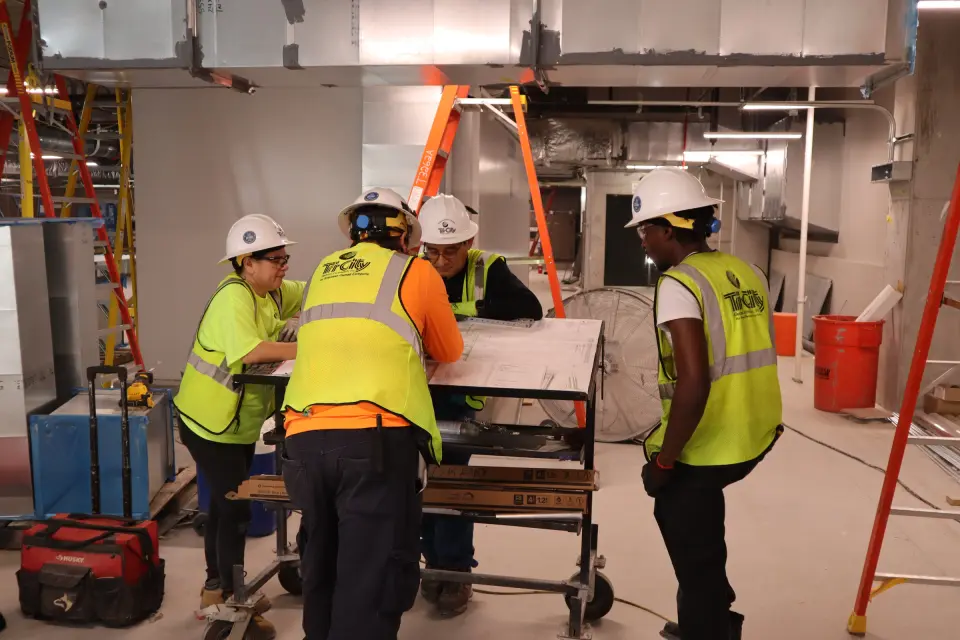 Tri-City Electrical Contractors Four construction workers wearing hard hats and reflective vests review blueprints on a cart in a building under construction. The room is filled with tools and ventilation ducts, and a ladder is visible in the background.