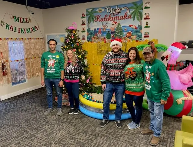 Tri-City Electrical Contractors A group of five people in festive sweaters and a Santa hat stand in front of a decorated Christmas tree. The room has Hawaiian-themed holiday decorations, including a banner that reads Mele Kalikimaka and a large inflatable flamingo.