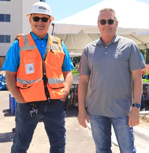 Tri-City Electrical Contractors Two men smiling outdoors at a construction site. One wears an orange safety vest and helmet, the other a gray polo shirt. They stand in front of a white tent, with a building and clear blue sky in the background.