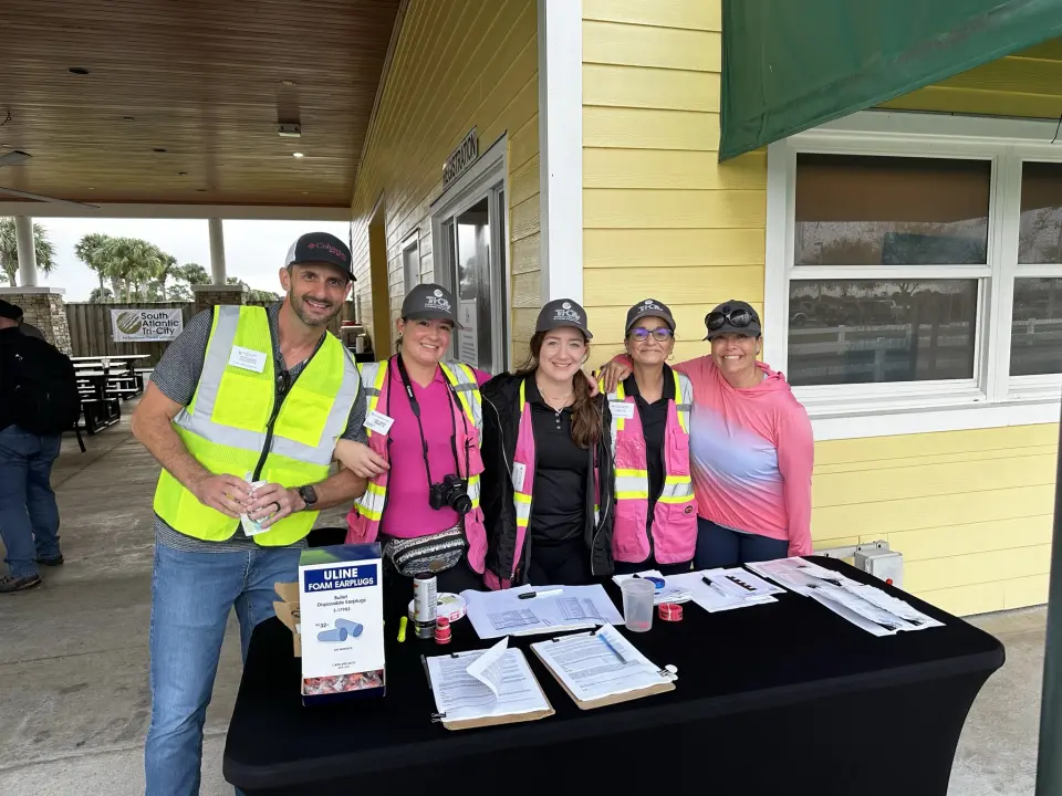 Tri-City Electrical Contractors Five people in safety vests and hats stand behind a table with papers and supplies. They are under a canopy near a yellow building. The setting appears to be a volunteer or community event.