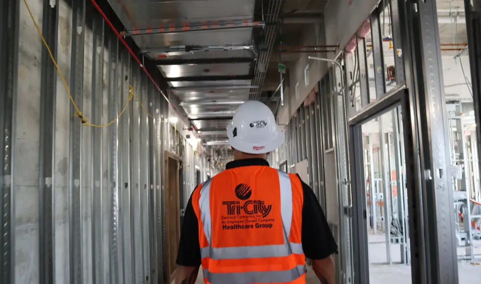 Tri-City Electrical Contractors Construction worker wearing a hard hat and a bright orange Tri-City vest walks down a corridor of an unfinished building, with exposed metal framing and wiring visible on the walls and ceiling.