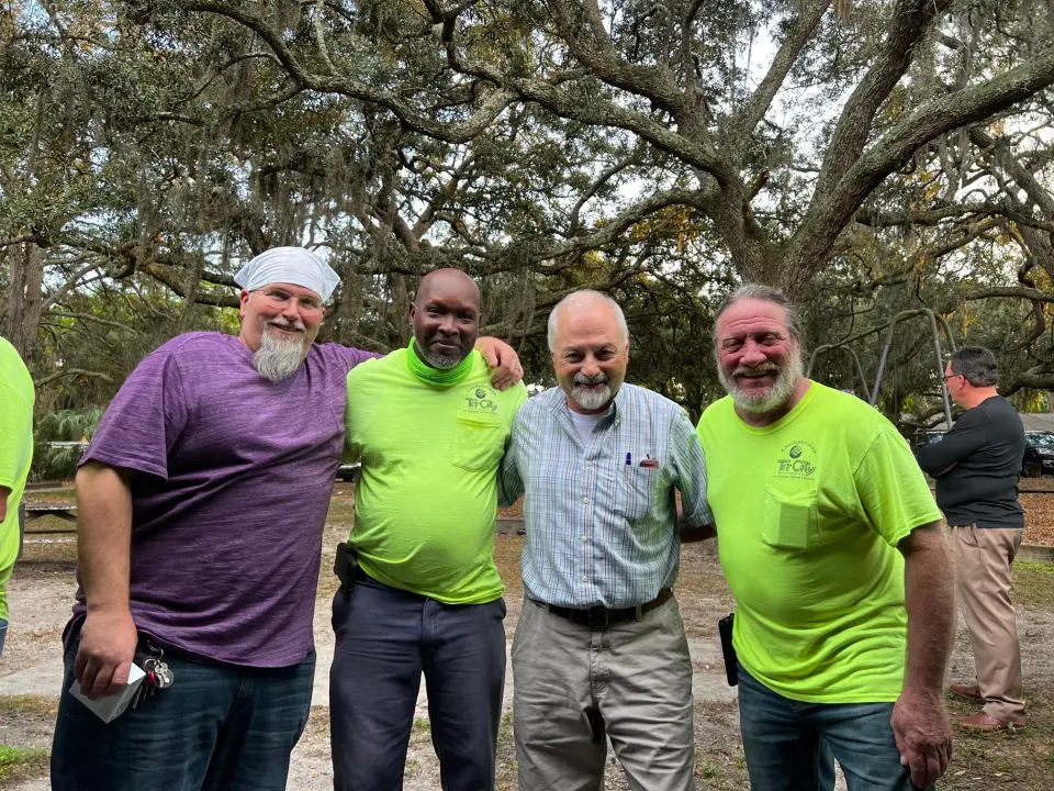 Tri-City Electrical Contractors Four men smiling outdoors, three wearing bright green shirts and one in a purple shirt with a white headscarf. They are standing under large trees with sprawling branches. Two other individuals are visible in the background.