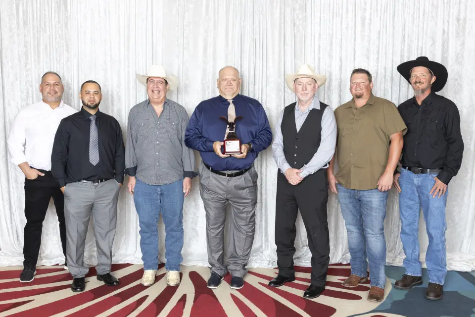 Seven men standing side by side in front of a white curtain, with the man in the center holding an award. Some are wearing cowboy hats and casual attire, while others are in dress shirts and ties.