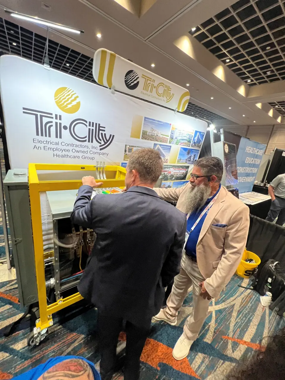 Tri-City Electrical Contractors Two men stand at a booth with a Tri-City Electrical Contractors Inc. banner at the 2024 AHCA EVENT - Healthcare Shows Up & Shows Out. One man gestures toward equipment, while the other observes, set against the backdrop of a bustling trade show or conference.