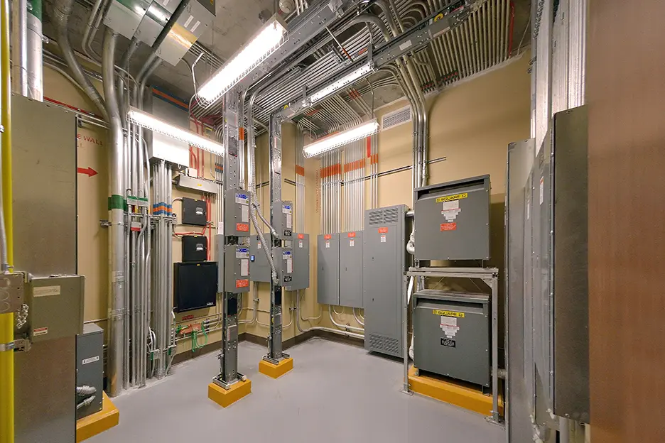 Tri-City Electrical Contractors A well-organized electrical room featuring various panels, conduits, and gray metal boxes. The space is well-lit with fluorescent lights, and the floor is clean and smooth. Labels are visible on the equipment for identification.