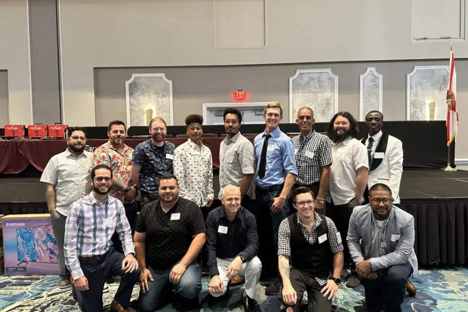 Tri-City Electrical Contractors A group of fifteen people posing for a group photo in a conference room. They are smiling and dressed in business casual attire. A table with red gift bags and a stage are visible in the background.