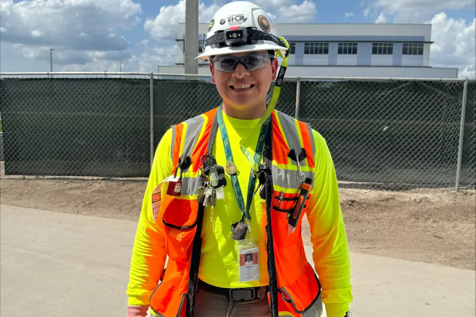 Tri-City Electrical Contractors A construction worker wearing a white helmet, orange safety vest, and yellow long-sleeve shirt stands on a paved area. The background features a chain-link fence and a building under a partly cloudy sky.