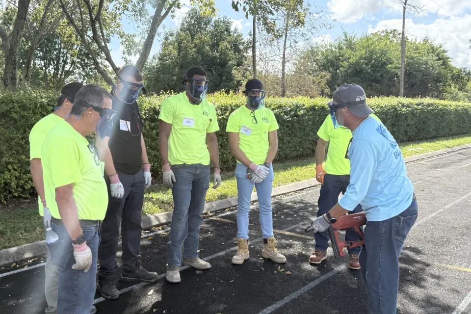 A group of people wearing safety goggles, gloves, and neon shirts stand outdoors on a paved area, attentively watching an instructor demonstrate a power tool. Trees and bushes line the background.