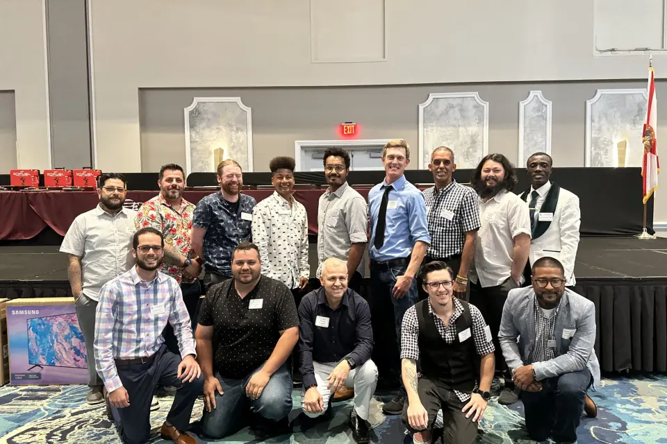 Tri-City Electrical Contractors A group of fifteen people posing together in a conference room. Some are kneeling, while others stand in two rows, with a display screen and stage in the background. A Florida state flag is visible on the stage.
