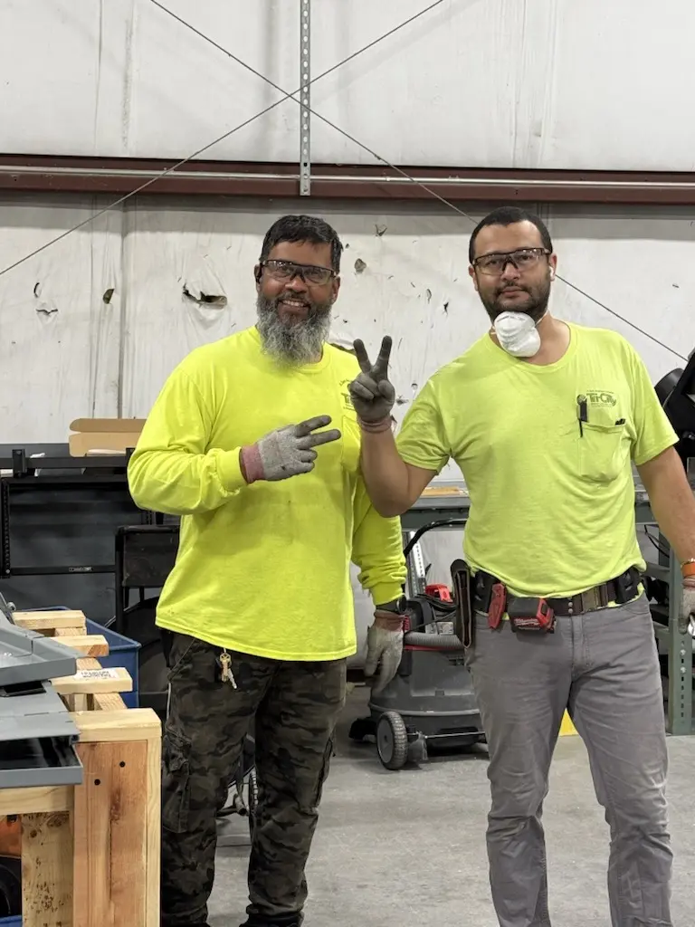 Tri-City Electrical Contractors Two men in lime green shirts stand in a prefabrication workshop. One wears camouflage pants, and the other sports a tool belt and face mask under his chin. Both are making peace signs and smiling, with shelves and various tools filling the background.