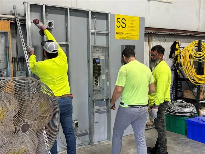 Tri-City Electrical Contractors Three workers in bright yellow shirts install prefabricated electrical components on a large panel. One uses a power tool, while the others observe. The area is organized with a 5S sign, and a large fan and coiled cables are visible nearby.
