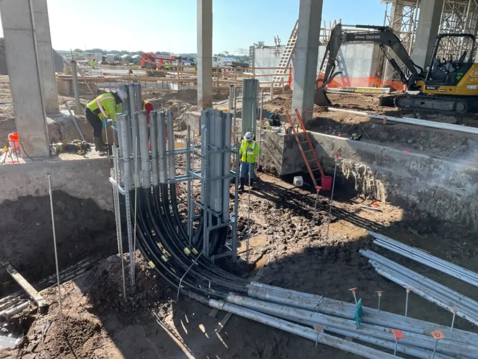 Tri-City Electrical Contractors At this bustling construction site, workers in safety gear are installing vertical metal pipes with precision. Leveraging prefabrication techniques, they ensure efficiency as an excavator looms in the background. The muddy ground is dotted with scaffolding and scattered materials.