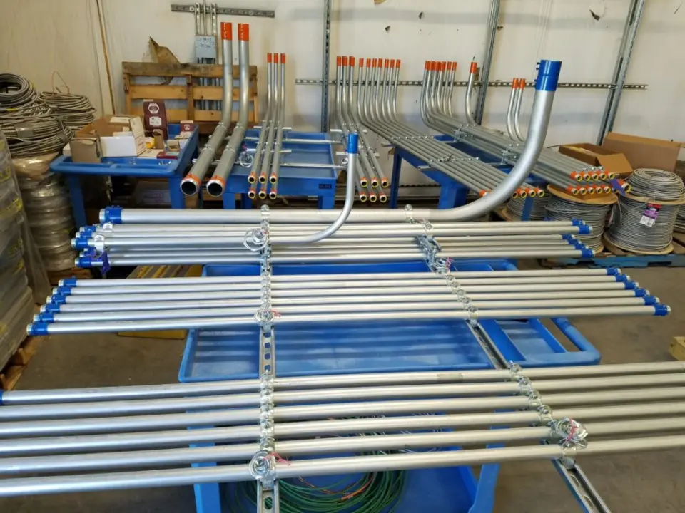 Tri-City Electrical Contractors Stacks of metal pipes and conduits are organized on blue carts in a warehouse. Boxes and coiled wires are visible in the background, with some pipes having colored caps for identification.