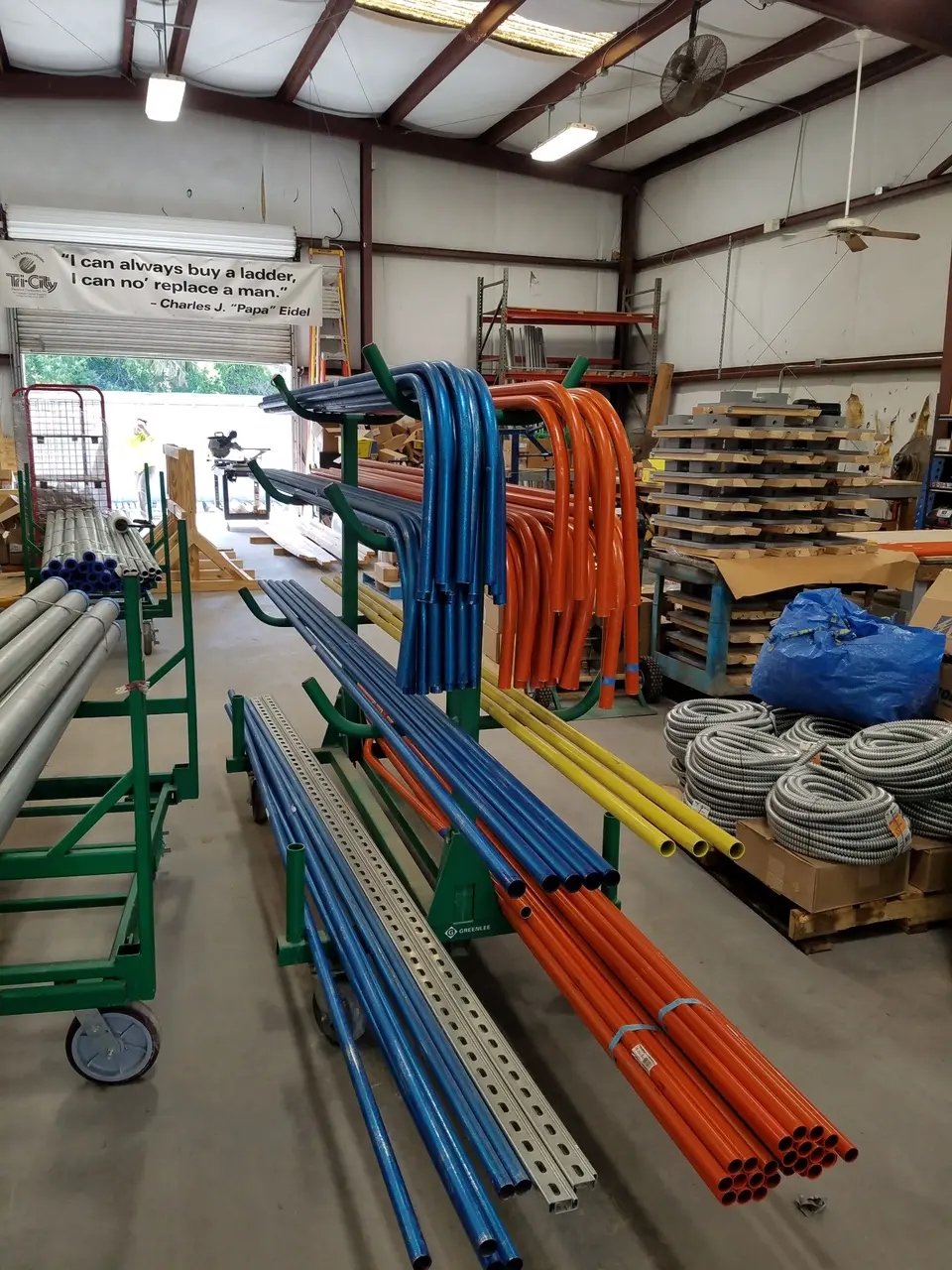 Tri-City Electrical Contractors A warehouse interior showcases prefabrication with various colored metal pipes and coiled cables stored on racks. The pipes are blue, orange, and gray, with several pallets of stacked materials in the background. Overhead lighting illuminates the space.