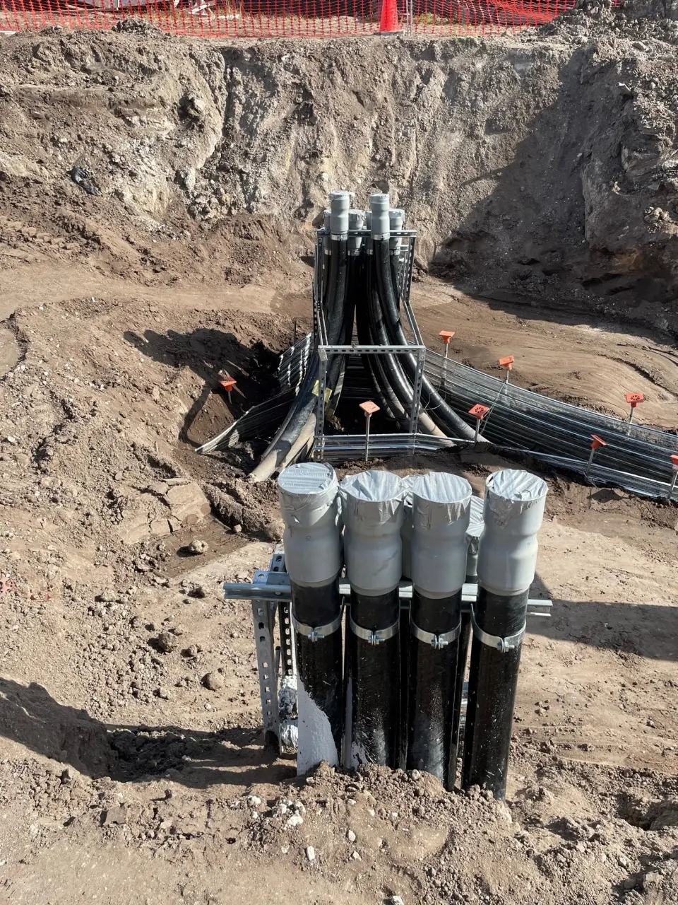 Tri-City Electrical Contractors The underground construction site features large black pipes, skillfully prefabricated, emerging from the ground and connected to sturdy metal supports. They are surrounded by exposed soil and orange safety markers in the background, ensuring a blend of efficient design and safety.