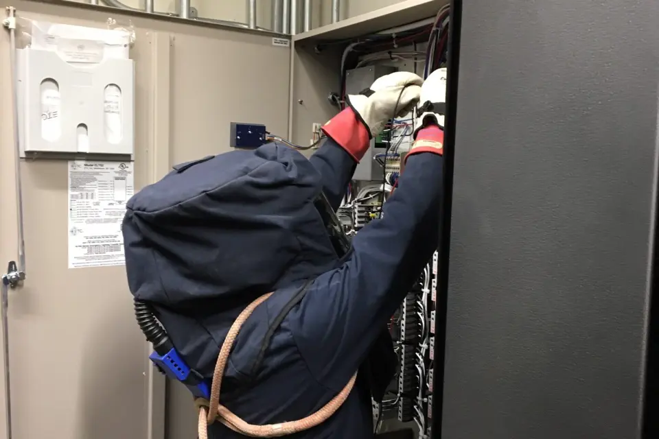 Person wearing electrical arc flash protective suit and gloves performing testing inside an open electrical control panel with exposed wiring.